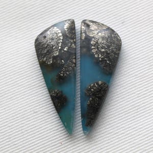 Pode incluir: Dois cabochões de pedras preciosas combinando. Cada cabochão tem formato triangular com a parte superior arredondada. As pedras apresentam uma base azul-turquesa com inclusões prateadas e pretas. Os cabochões são exibidos em uma superfície branca texturizada.