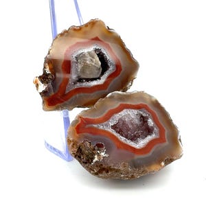 May include: Two halves of a geode, displaying a natural, earthy appearance. The outer layers are a mix of tan and brown, with vibrant red and white bands encircling the crystal-filled centres. The crystals are a mix of clear and purple.
