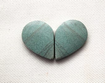 Puede incluir: Dos piedras en forma de lágrima de color turquesa, con una superficie texturizada y vetas de color gris oscuro. Las piedras están colocadas una al lado de la otra, creando una forma de corazón. El fondo es una tela blanca texturizada.