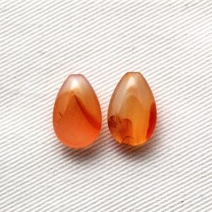 Peut inclure: Deux perles en forme de goutte d'eau. Les perles sont d'une couleur orange translucide avec des stries orange foncé et blanches. Les perles sont lisses et polies, et semblent être faites d'une pierre ou d'un matériau en verre. Le fond est un tissu blanc texturé.