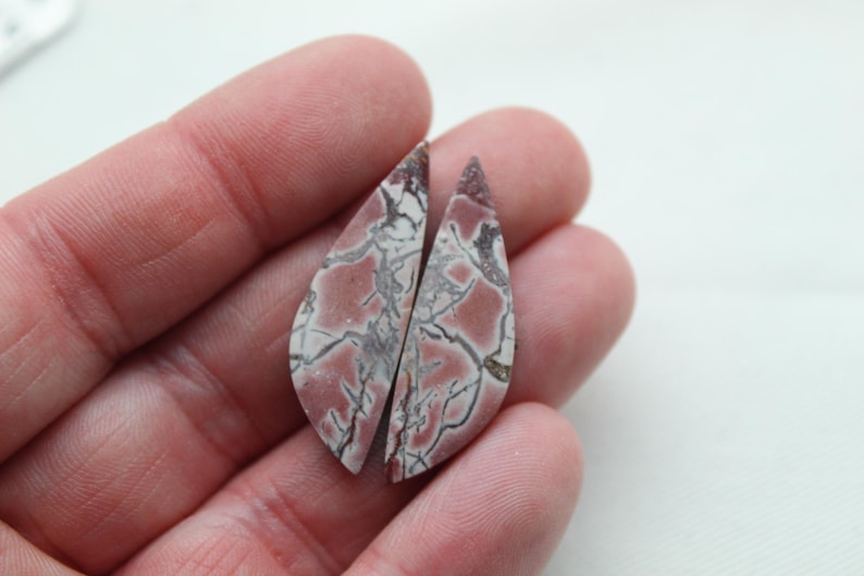 May include: Two teardrop-shaped cabochons featuring a dusty rose and cream-colored base with a complex network of dark grey and black veining throughout the surface. These polished stone pieces have a smooth, flat back and a curved front surface.