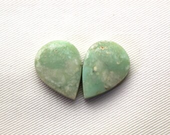 May include: Two teardrop-shaped gemstones with a pale green hue and white marbling. The stones are set against a white, textured background. These polished stones are likely used for jewelry making or other crafts.