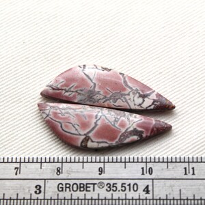 May include: Two polished, teardrop-shaped cabochons featuring a dusty rose and white base with dark, branching vein patterns. A metal ruler is positioned below the stones, showing they measure approximately 1.5 inches in total length.