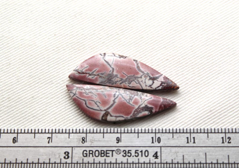 May include: Two polished, teardrop-shaped cabochons featuring a dusty rose and white base with dark, branching vein patterns. A metal ruler is positioned below the stones, showing they measure approximately 1.5 inches in total length.