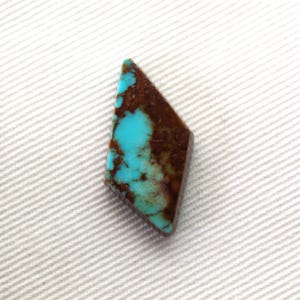 Puede incluir: Una piedra preciosa turquesa en forma de diamante. La piedra presenta un vibrante color azul-verdoso con patrones de matriz marrón. La gema se encuentra sobre un fondo blanco texturizado.