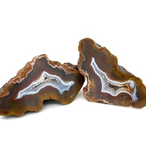 May include: Two polished agate stone slices. The stones feature concentric bands of brown, red, white, and grey, with a crystalline centre. The rough, natural edges contrast with the smooth, polished interior. These decorative stones are ideal for home decor.