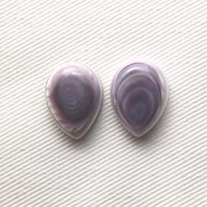 Könnte beinhalten: Zwei tropfenförmige Cabochons mit einer glatten, polierten Oberfläche und konzentrischen Bändern in Lavendel-, Violett- und Grautönen. Diese Steine weisen ein glänzendes Finish und eine symmetrische, abgerundete Spitze auf.
