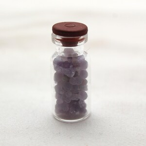 Peut inclure: Un petit flacon en verre transparent rempli de petits objets violets arrondis. Le flacon est coiffé d'un bouchon marron foncé. Les objets à l'intérieur ressemblent à de petits cailloux ou perles. Le fond est une surface blanche unie.