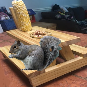 May include: A gray squirrel sits on a miniature wooden picnic table with a cob of corn and a bowl of nuts. The table is made of unfinished wood and has a small bench on each side.