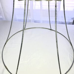 May include: A vintage, silver-toned lampshade frame. The frame is made of metal wire, with a wide bottom ring and a smaller top ring. Vertical wires connect the rings, creating a bell-like shape. The frame is designed to hold fabric or other materials for a lampshade.