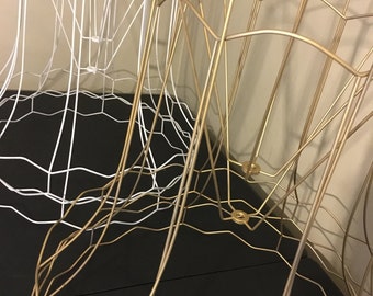 May include: Two lampshades, one white and one gold, are made of wire and are stacked on top of each other. The lampshades are both in the shape of a bell.