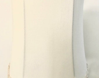 May include: A white lampshade with a decorative trim around the top and bottom. The lampshade is attached to a gold metal lamp base.