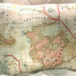May include: A vintage-style map pillow featuring a faded, light blue background with a pink and brown outline of North America. The map includes the text "GREENLAND" and "NORTH AMERICA" with a red line outlining the Atlantic Ocean.
