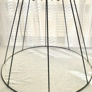 May include: A black metal lampshade frame with a round base and a conical shape. The frame is made of thin metal rods that are joined together at the top and bottom.