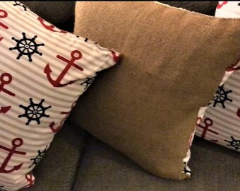 May include: Two decorative throw pillows. One pillow is brown and the other is white with red anchors and black steering wheels on a striped background.
