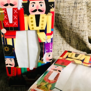 May include: Two tissue boxes with a Nutcracker theme. The boxes are red, blue, green, and yellow with a white tissue opening. The Nutcrackers are wearing red, blue, and yellow uniforms.