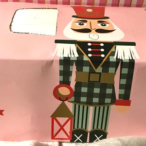 May include: A pink box with illustrations of nutcracker soldiers. One soldier is wearing a green and black plaid uniform, holding a lantern. The other soldier is wearing a green uniform. Red stars are scattered across the box.