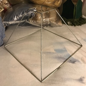 May include: A silver metal lampshade frame in the shape of a pyramid. The frame is made of thin metal rods that are joined together at the corners. The frame is sitting on a white surface.