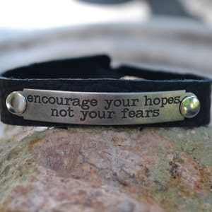 May include: Black leather bracelet with a silver metal plate that reads "encourage your hopes not your fears".