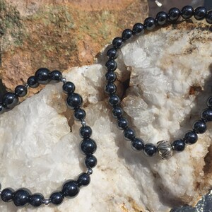 May include: Two black hematite beaded bracelets with silver accents. The bracelets are on a white background.