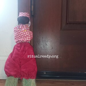 May include: A red and white checkered doll with a black head and a red skirt. The doll is standing on a broom with green bristles. The doll is leaning against a wooden door with a metal door handle. The text "ritualready.org" is visible on the door.