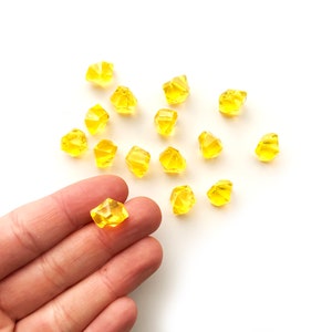 May include: A collection of small, faceted, yellow gemstone beads scattered on a white surface. One bead is held in a hand. The beads have a crystalline appearance and a bright, translucent color, suggesting they are made of glass or a similar material.