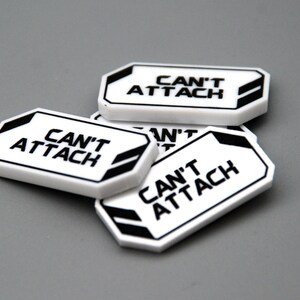 May include: A collection of white rectangular plastic tags with beveled corners, each featuring the black text CAN'T ATTACH printed in the center. The tags have a black border and black geometric accents on the left and right sides.