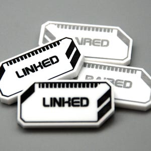 May include: A collection of four white rectangular tokens with beveled corners, featuring black graphic accents and the text LINKED or PAIRED printed in a bold, sans-serif font. Each token has a small grid pattern along the top edge.