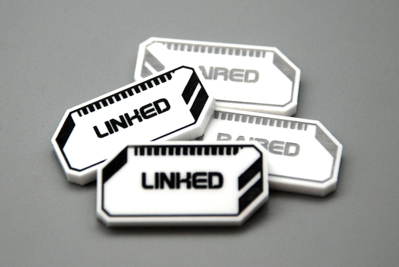 May include: A collection of four white rectangular tokens with beveled corners, featuring black graphic accents and the text LINKED or PAIRED printed in a bold, sans-serif font. Each token has a small grid pattern along the top edge.