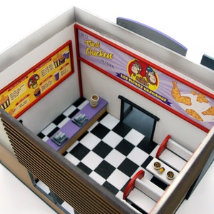 K&ouml;nnte beinhalten: Ein Miniaturmodell eines Fast-Food-Restaurant-Innenraums mit einem schwarz-wei&szlig; karierten Boden, einer violetten Theke und roten Sitzb&auml;nken. Die W&auml;nde sind mit Schildern verziert, auf denen "Best Fried Chicken in Town" und "Los Monos Hermanas" steht.