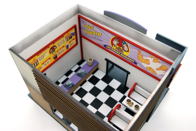 K&ouml;nnte beinhalten: Ein Miniaturmodell eines Fast-Food-Restaurant-Innenraums mit einem schwarz-wei&szlig; karierten Boden, einer violetten Theke und roten Sitzb&auml;nken. Die W&auml;nde sind mit Schildern verziert, auf denen "Best Fried Chicken in Town" und "Los Monos Hermanas" steht.