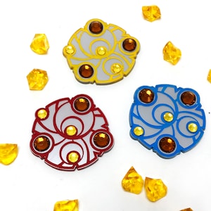 May include: Three decorative metal pins with a circular design. Each pin has a different color: gold, red, and blue. The pins have a white background with a swirling pattern and are decorated with small, clear rhinestones.