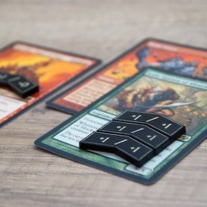 Könnte beinhalten: Nahaufnahme einer grün-schwarzen Magic: The Gathering Karte mit einem schwarzen und goldenen Zähler oben drauf. Die Karte zeigt eine Kreatur mit einem Schwert und einem Schild. Der Text auf der Karte lautet "Legendäre Kreatur - Menschlicher Krieger".