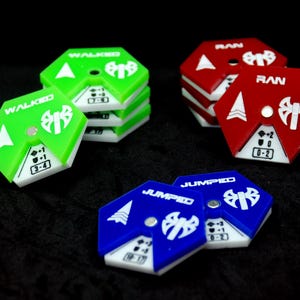 May include: Hexagonal game tokens in green, red, and blue. Each token has white symbols and text indicating actions like "WALKED", "RAN", and "JUMPED". The tokens are stacked and scattered on a black surface.