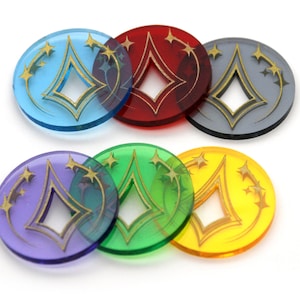 Puede incluir: Seis fichas redondas de acrílico translúcido con una forma de diamante dorado y estrellas en el fondo. Las fichas son de color azul, rojo, gris, morado, verde y amarillo.