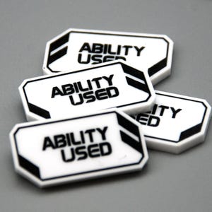 May include: A collection of four white rectangular tokens with beveled corners, each featuring the black text "ABILITY USED" printed in a bold, sans-serif font. The tokens have black geometric accents on the right side and a black border.
