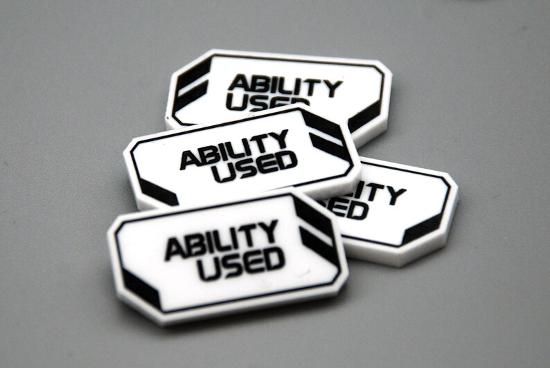 May include: A collection of four white rectangular tokens with beveled corners, each featuring the black text "ABILITY USED" printed in a bold, sans-serif font. The tokens have black geometric accents on the right side and a black border.