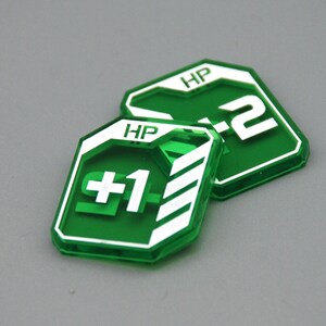 May include: Two translucent green acrylic game tokens with white printed details. The front token displays the text HP and +1 with white diagonal stripes. The token behind it displays the text HP and +2. Both tokens have a shield-like shape.