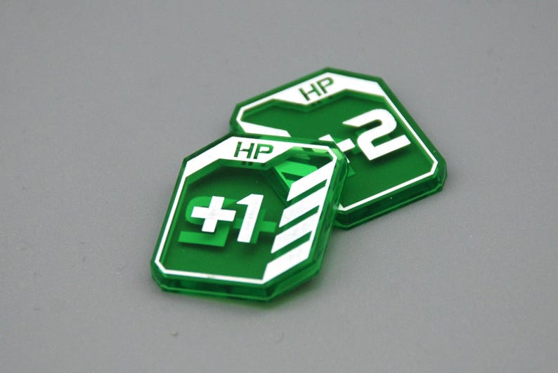 May include: Two translucent green acrylic game tokens with white printed details. The front token displays the text HP and +1 with white diagonal stripes. The token behind it displays the text HP and +2. Both tokens have a shield-like shape.