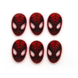 Puede incluir: Seis botones de plástico rojo y negro con la cara de Spider-Man. Cada botón presenta un contorno negro de la cara de Spider-Man sobre un fondo rojo.