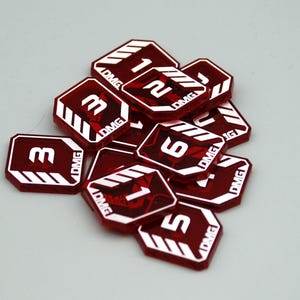 May include: A collection of translucent red acrylic tokens, each octagonal in shape with white printed numbers and the text DMG. The tokens feature white diagonal stripes in the corners and display various numerical values including 1, 2, 3, 5, and 6.