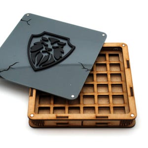 Può includere: Una scatola di legno con un interno a griglia e un coperchio grigio. Il coperchio presenta un emblema nero e crepe decorative. La scatola è progettata per organizzare piccoli oggetti.