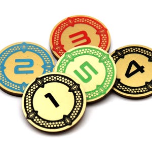 Op de afbeelding: Vijf ronde spel fiches met de nummers 1 tot en met 5. Elke fiches heeft een goudkleurig midden met een gekleurde rand. De randen zijn blauw, rood, groen en zwart, op een witte achtergrond.