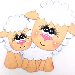 May include: Two cartoon sheep cutouts, approximately 4 inches tall and 4.5 inches wide, with white fluffy wool, peach-colored faces, and pink noses. The sheep have large black eyes with long eyelashes and are smiling. The text "Scrappin' Heaven" is at the bottom.