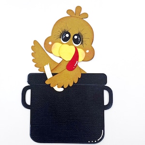 May include: A papercraft turkey peeking out of a black pot, with the text "Scrappin' Heaven" below. The turkey is brown, yellow, and red, approximately 3.25 inches wide and 5 inches tall.