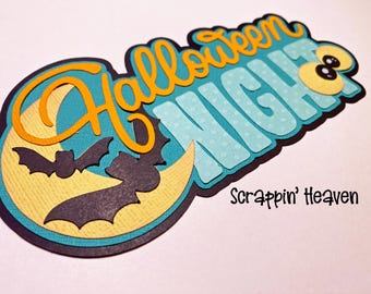 May include: A layered paper die-cut decoration featuring the text Halloween Night in orange and light blue with white polka dots. The design includes a yellow crescent moon, two black bats, and a pair of cartoon eyes, with Scrappin' Heaven text.