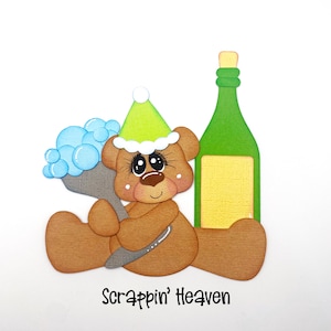 May include: A brown teddy bear illustration wearing a green party hat, holding a gray object with blue bubbles, and sitting next to a green bottle with a yellow label. The text "Scrappin' Heaven" is at the bottom.