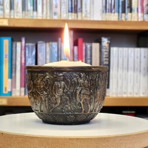 Puede incluir: Un candelero de bronce con un diseño intrincado de figuras y una vela encendida en su interior. El candelero está sobre una superficie de madera.