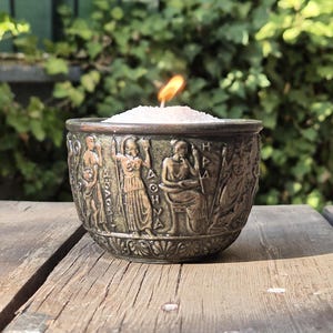 Op de afbeelding: Een bronzen kaarsenhouder met een witte kaars erin. De houder is versierd met een reli&euml;f van figuren en Griekse letters. De kaars is aangestoken en de vlam is zichtbaar.