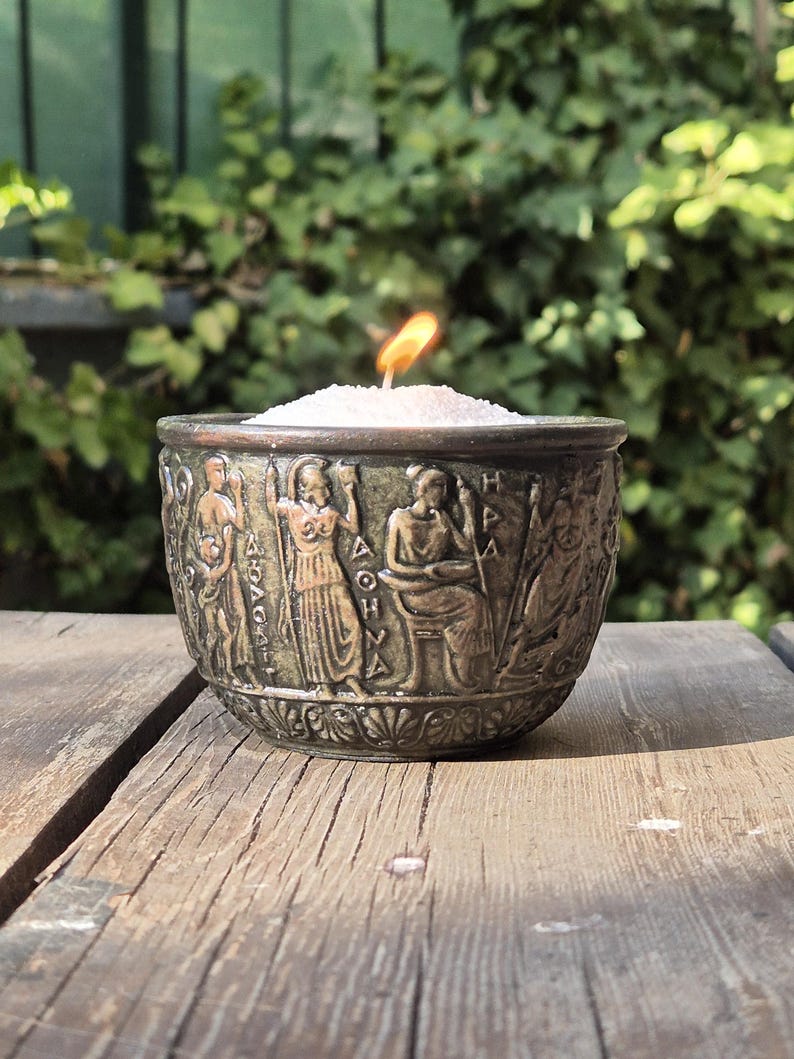Op de afbeelding: Een bronzen kaarsenhouder met een witte kaars erin. De houder is versierd met een reli&euml;f van figuren en Griekse letters. De kaars is aangestoken en de vlam is zichtbaar.